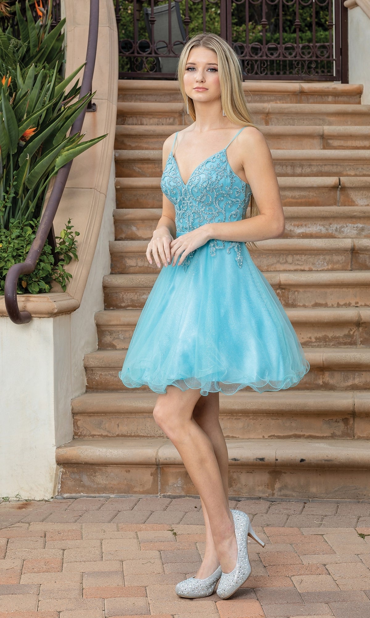 A young woman stands on stone steps in the Lace-Up-Back Short Babydoll Homecoming Dress 3315, featuring a beaded corset bodice, paired with sparkly high heels and lush green plants beside the stairs.